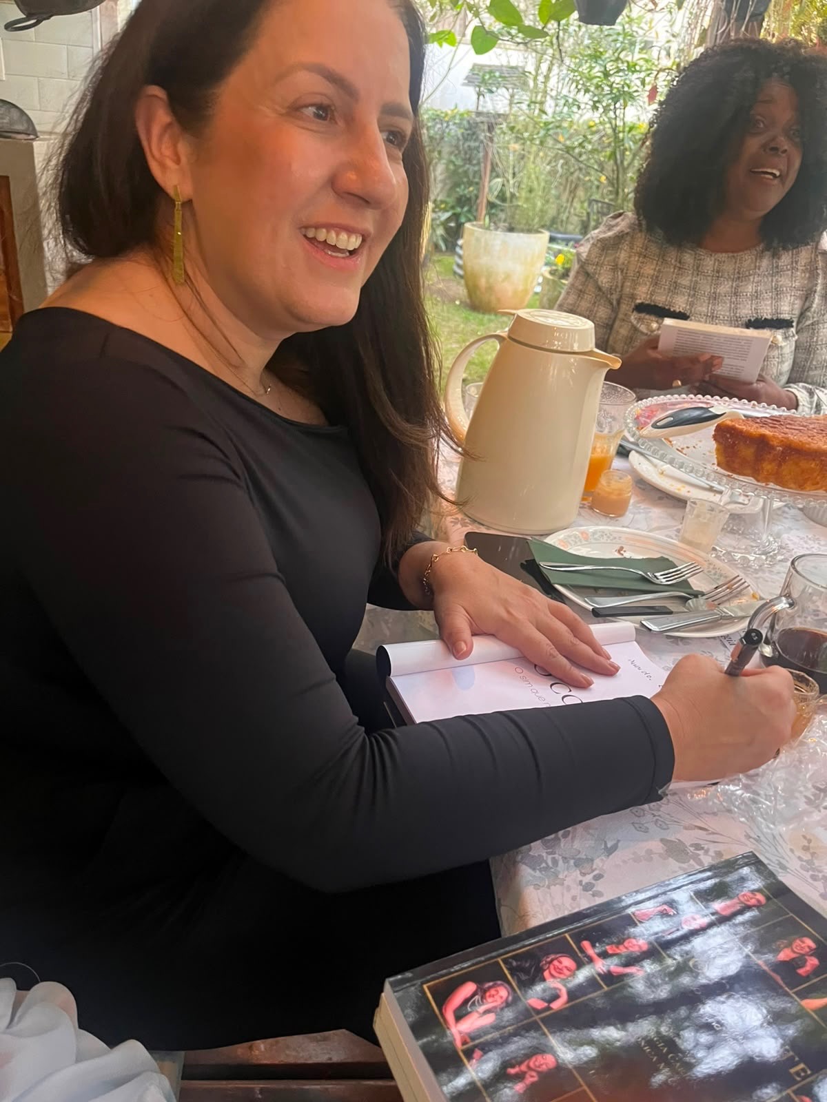 Convidada sorrindo enquanto escreve à mesa, com café e bolo